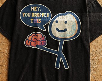May include: Black t-shirt with a humorous graphic. The design features a stick figure holding a brain, with a speech bubble that reads "HEY, YOU DROPPED THIS." The stick figure has a smiling face. The graphic has a vintage, distressed look.