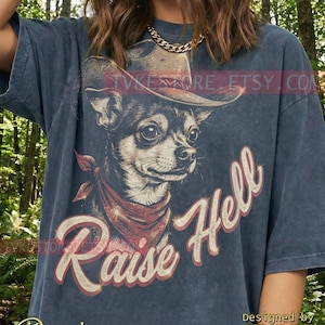 May include: Denim blue t-shirt featuring a graphic of a Chihuahua wearing a cowboy hat and bandana. The text "Raise Hell" is arched across the image. The shirt is designed by TVKE.