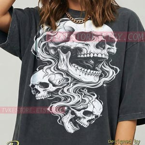 May include: A charcoal gray oversized t-shirt featuring a large graphic print of three stacked skulls surrounded by swirling smoke and sparkling accents. The text TVKESTORE.ETSY.COM is visible across the design, with the word Pepper and TVKE below.
