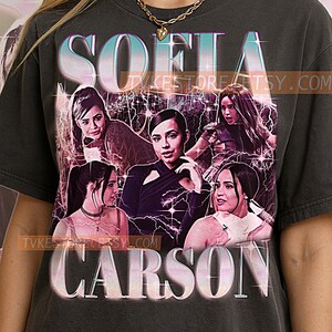 May include: Dark gray t-shirt featuring a graphic of Sofia Carson with the name "SOFIA CARSON" in a stylized font. The design includes multiple images of the actress with lightning bolt accents.