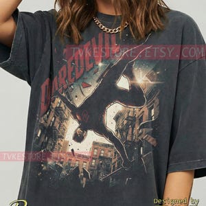 May include: Dark gray t-shirt featuring a graphic of Daredevil in action, with the word "DAREDEVIL" in red. The design includes a cityscape background and the text "TVKESTORE.ETSY.COM". The shirt is designed by TVKE.