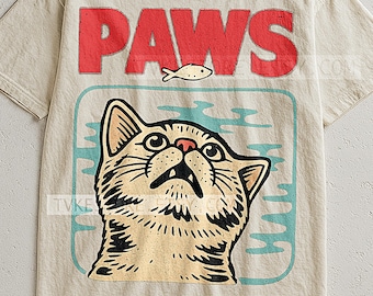 Op de afbeelding: Beige t-shirt met een grafisch ontwerp met een kat die naar een vis kijkt. Het woord "PAWS" is in het rood boven de kat gedrukt. De kat staat in een blauwe rechthoek met waterachtige vormen.