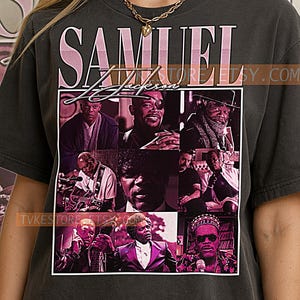 May include: Dark gray t-shirt featuring a collage of images of Samuel L. Jackson, with his name in pink and white lettering. The shirt has a crew neck and short sleeves. The design is a tribute to the actor.