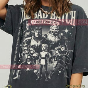 May include: Dark gray t-shirt featuring a graphic of characters from "The Bad Batch" with the text "The Bad Batch" and "Clone Force 99". The design includes the names of the characters. The shirt is designed by TVKE.