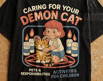 May include: Black t-shirt with a graphic featuring a girl hugging a cat with horns, surrounded by candles. Text reads "Caring for Your Demon Cat," "Pets & Responsibilities," and "Activities for Children."