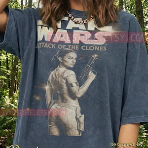 May include: Denim blue t-shirt featuring a graphic from Star Wars: Attack of the Clones. The design includes the movie title, a character holding a blaster, and the text "Designed by TVKE".