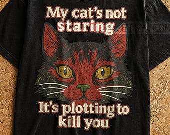 May include: Black t-shirt featuring a red and black cat face with yellow eyes. The text "My cat's not staring" is above the cat's head, and "It's plotting to kill you" is below.