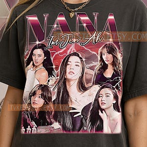 May include: Dark gray t-shirt featuring a graphic of the name "NANA" in a retro style, with the name "Im Jin Ah" below. The design includes images of several women. The shirt has a vintage aesthetic.
