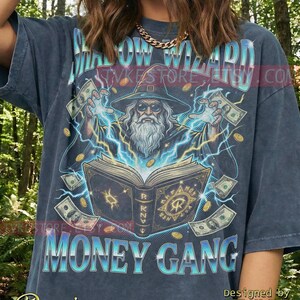 Puede incluir: Camiseta de mezclilla gris oscuro con un gráfico de un mago conjurando dinero de un libro, con el texto "SHADOW WIZARD MONEY GANG". El diseño incluye rayos, monedas y billetes. Diseñado por TVKE.