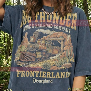 May include: Denim blue t-shirt with a retro graphic of a train exiting a tunnel, with text reading "Big Thunder Mine & Railroad Company Frontierland Disneyland." The design incorporates cacti and desert landscape elements.