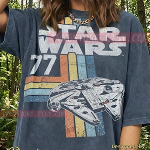 Può includere: T-shirt blu denim con grafica del Millennium Falcon e la scritta "STAR WARS 77". Il design include strisce retrò arancioni, gialle e blu. La maglietta è disegnata da TVKE.