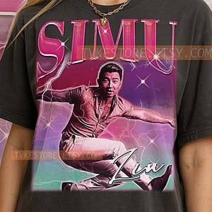以下が含まれることがあります： ヴィンテージスタイルのグラフィックが特徴の濃いグレーのTシャツ。Simu Liuのデザインで、「SIMU」のピンク色の文字、稲妻、筆記体の「Liu」という名前が特徴です。Tシャツは人が着ています。