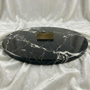 May include: A circular black marble serving board featuring prominent white veining throughout the stone surface. A small, rectangular brass-colored metal plaque is centered on the board, engraved with the text STONE HERITAGE ART.