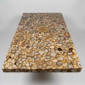 Puede incluir: Una mesa rectangular con una superficie hecha de piedras pulidas de forma irregular en tonos marrones, beige y grises. Un peque&ntilde;o letrero de lat&oacute;n que dice "STONE HERITAGE ART" est&aacute; fijado a la superficie de la mesa.