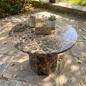 May include: A round brown marble table with a hexagonal base featuring white veining. On the surface sits a beige ceramic mug, a small green succulent in a light gray pot, and a rectangular wooden sign with the text Stone Heritage Art.