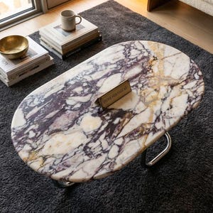 May include: Oval marble coffee table with a white, purple, and gold veined pattern. The table has a polished metal base and a small brass sign that reads "STONE HERITAGE ART". A gold bowl and books are visible.