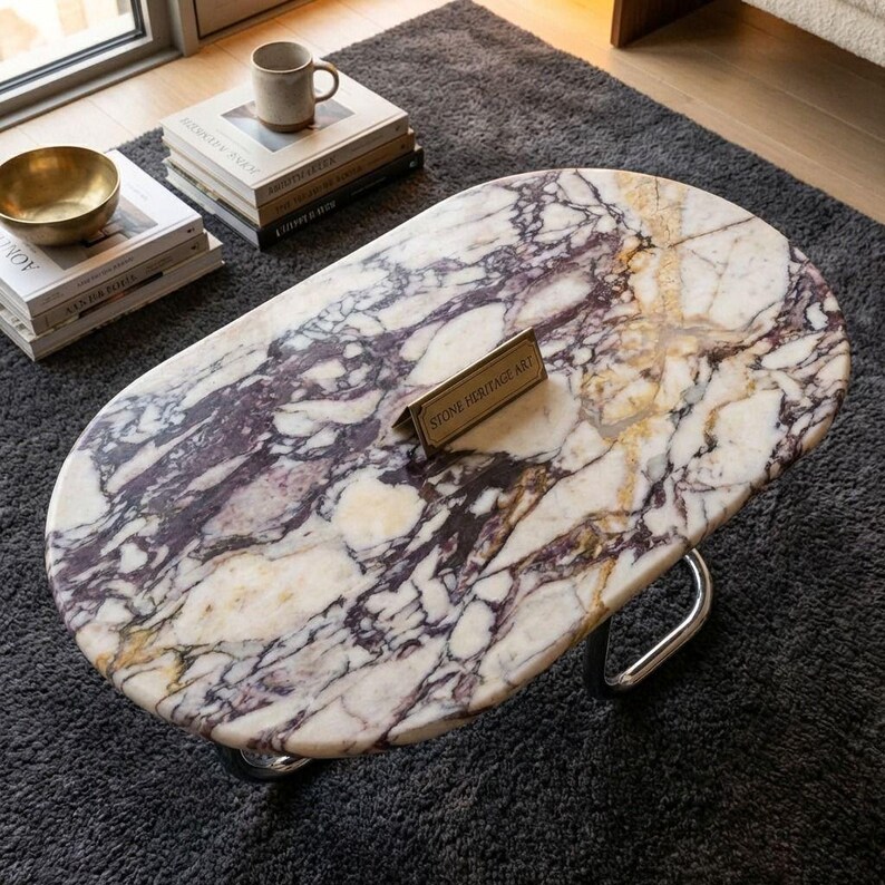 May include: Oval marble coffee table with a white, purple, and gold veined pattern. The table has a polished metal base and a small brass sign that reads "STONE HERITAGE ART". A gold bowl and books are visible.
