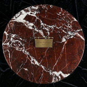 May include: Circular red marble surface with white veining. A small brass sign with the text "STONE HERITAGE ART" is attached to the surface. The marble has a polished finish and is set against a dark background.