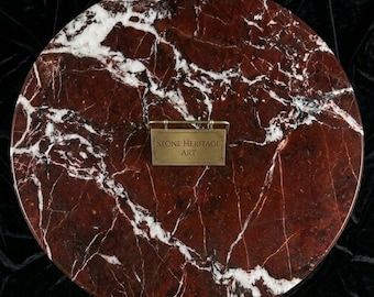 Puede incluir: Superficie circular de mármol rojo con vetas blancas. Una pequeña placa de latón con el texto "STONE HERITAGE ART" está adherida a la superficie. El mármol tiene un acabado pulido, sobre un fondo oscuro.