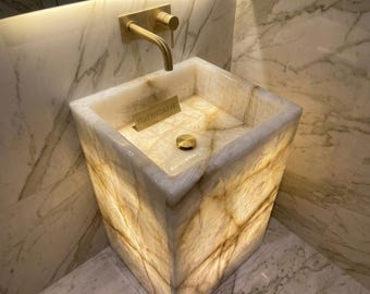May include: A square, translucent white and gold onyx stone pedestal sink with a polished brass wall-mounted faucet and drain. A small rectangular plaque inside the basin features the text STONE HERITAGE ART in gold lettering.