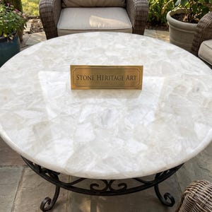 May include: A round coffee table with a white, translucent, crystalline stone top featuring a mosaic-like texture. The table has a black metal base with curved legs. A rectangular brass plaque sits in the center with the text Stone Heritage Art.