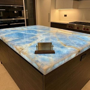 May include: A rectangular kitchen island countertop made of translucent blue onyx stone with white veining, illuminated from beneath. A small rectangular metal plaque with a dark wooden base sits on the surface, displaying the text STONE HERITAGE ART.