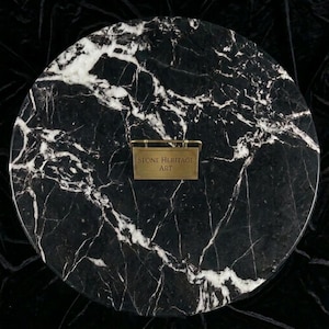 May include: A circular black marble slab featuring prominent white veining throughout the surface. Centered on the stone is a small, rectangular brass-toned metal plaque with the engraved text "STONE HERITAGE ART" in a clean, sans-serif font.