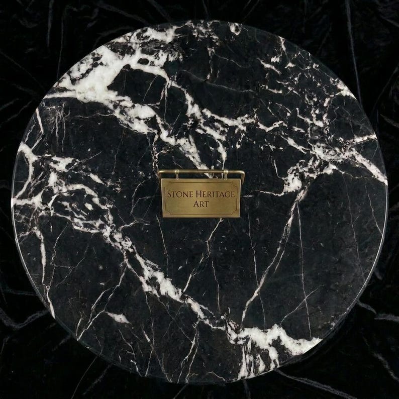 May include: A circular black marble slab featuring prominent white veining throughout the surface. Centered on the stone is a small, rectangular brass-toned metal plaque with the engraved text "STONE HERITAGE ART" in a clean, sans-serif font.