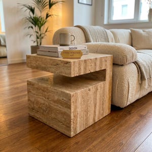 May include: A beige travertine side table with a C-shaped design. On top of the table are two stacked books, one titled DESIGN and the other with a gold plaque reading STONE HERITAGE ART, and a light-colored ceramic mug with a handle.