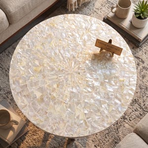 May include: A round table featuring a surface covered in a mosaic of iridescent white mother-of-pearl shell fragments arranged in a radial pattern. A small rectangular wooden sign with the text STONE HERITAGE ART sits on the table surface.
