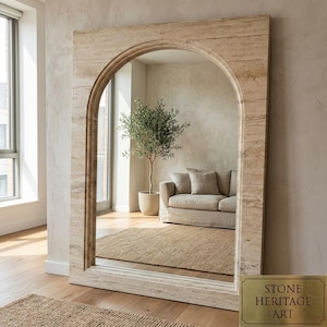 May include: A large rectangular floor mirror with an arched top, featuring a beige and tan travertine stone frame with natural horizontal veining. A small rectangular brass plaque in the bottom right corner reads STONE HERITAGE ART.