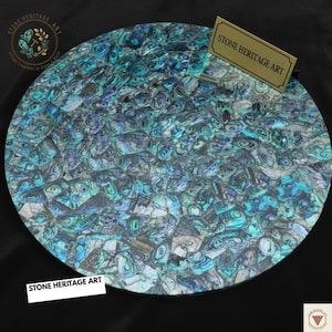 May include: A circular decorative item with a mosaic pattern. It features iridescent blue, green, and grey pieces, producing a shimmering effect. The text "STONE HERITAGE ART" is displayed on a small gold sign and a white label.