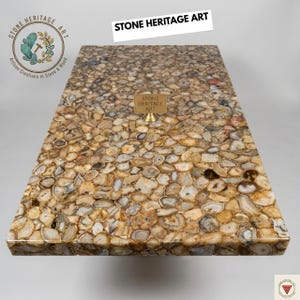 Puede incluir: Una mesa rectangular con una superficie de mosaico hecha de piedras pulidas de forma irregular en tonos marrones, beige y grises. La mesa tiene un acabado brillante y presenta el texto "STONE HERITAGE ART".