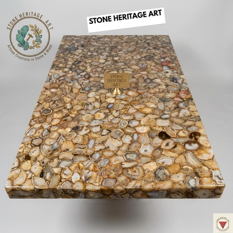Puede incluir: Una mesa rectangular con una superficie de mosaico hecha de piedras pulidas de forma irregular en tonos marrones, beige y grises. La mesa tiene un acabado brillante y presenta el texto "STONE HERITAGE ART".