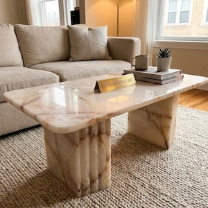 May include: A rectangular coffee table crafted from polished cream-coloured stone with prominent brown veining. The table features a smooth, thick tabletop supported by two sturdy, vertically ribbed block legs. A gold nameplate reads STONE HERITAGE ART.