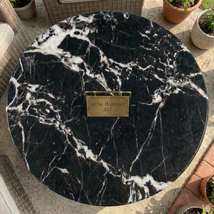 May include: A circular black marble table top featuring prominent white veining throughout the stone surface. Centered on the table is a rectangular brass-colored metal plaque with the engraved text Stone Heritage Art in a serif font.