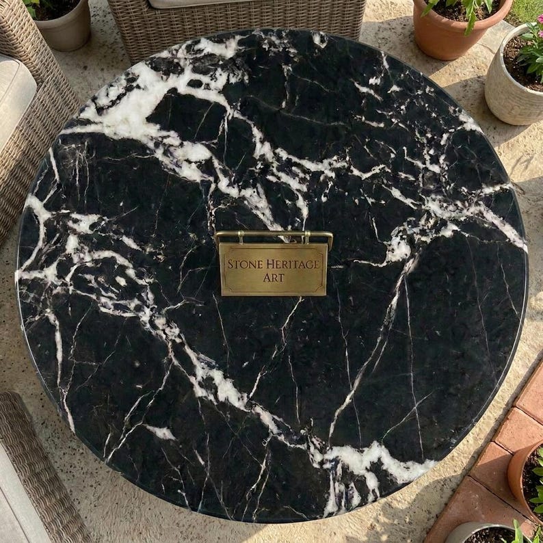 May include: A circular black marble table top featuring prominent white veining throughout the stone surface. Centered on the table is a rectangular brass-colored metal plaque with the engraved text Stone Heritage Art in a serif font.
