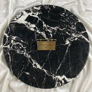 May include: A circular black marble slab featuring prominent white veining throughout the surface. Centered on the slab is a rectangular brass-colored metal plaque with the engraved text "STONE HERITAGE ART" in a clean, sans-serif font.