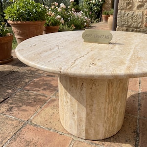 May include: A round beige travertine stone table with a smooth top and a cylindrical, vertically fluted base. A small rectangular brass-colored plaque with the text STONE HERITAGE ART is placed on the surface of the table.