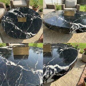 May include: A round black marble table with prominent white veining throughout the surface. A small rectangular brass plaque is placed on the tabletop, featuring the engraved text "STONE HERITAGE ART" in a clear, capitalized font.