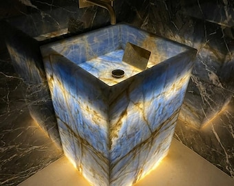 May include: A square, floor-standing pedestal sink crafted from translucent blue and gold veined stone. The basin is internally illuminated, highlighting the natural patterns. A gold-toned faucet is mounted above, and a small plaque reads Stone Heritage Art.