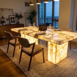 May include: A rectangular dining table with a backlit translucent white and amber stone top and base, featuring prominent natural veining. The table is set with clear glassware and napkins, and a small sign reads STONE HERITAGE ART.