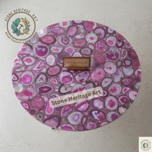 Puede incluir: Una mesa redonda con un diseño de mosaico de piedras de ágata rosas y blancas. La mesa tiene una placa de madera que dice "STONE HERITAGE ART". Las palabras "Stone Heritage Art" también están impresas en la mesa. Un artículo decorativo.