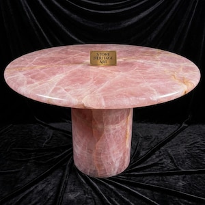 May include: A round, pink rose quartz table with a cylindrical base. The table's surface has a polished finish with natural veining. A small, rectangular plaque with the text "STONE HERITAGE ART" is on the table's surface.
