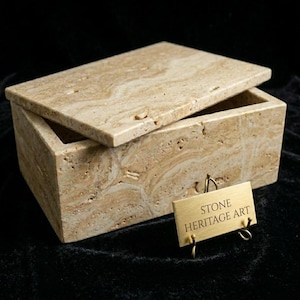 May include: A rectangular beige travertine stone box with a matching lid. The stone features natural porous textures and swirling cream and tan patterns. A small gold-toned metal plaque stands in front, engraved with the text STONE HERITAGE ART.