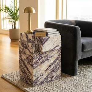 Può includere: Un tavolino rettangolare realizzato in marmo bianco con venature viola prominenti, caratterizzato da un design a blocchi sfalsati. Una lampada a forma di fungo color ottone e una piccola pila di libri poggiano sulla superficie del tavolo.