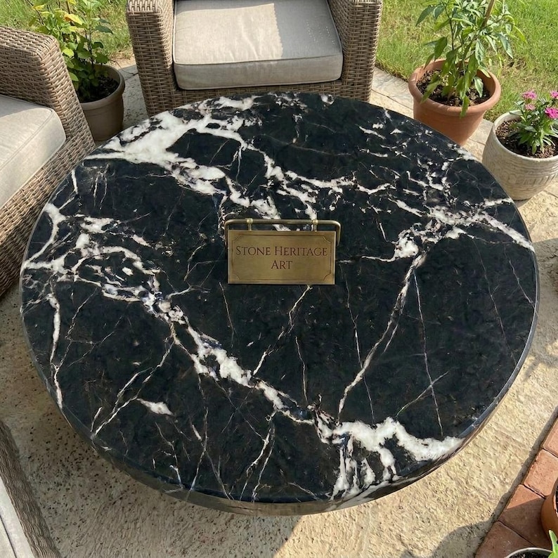 May include: A round table with a polished black marble top featuring prominent white veining. Centered on the surface is a small rectangular brass plaque with the engraved text "STONE HERITAGE ART" held by a thin metal frame stand.