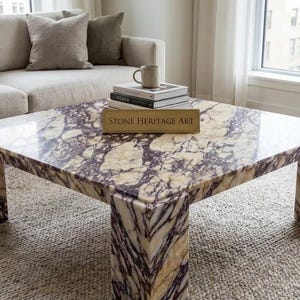 Può includere: Un tavolino quadrato realizzato in pietra lucida con un motivo venato color crema e viola scuro. Il tavolo presenta quattro gambe spesse a forma di blocco e una superficie piana. Un cartello rettangolare con il testo STONE HERITAGE ART è posto sopra.