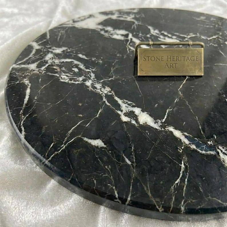 May include: A circular black marble slab featuring prominent white veining throughout the surface. A rectangular brass-colored metal plaque is attached to the top right, engraved with the text STONE HERITAGE ART in a clean, sans-serif font.