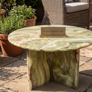 May include: A round side table crafted from light green onyx stone with natural veining and a polished finish. The table features a sturdy base made of two intersecting vertical panels. A rectangular plaque with the text STONE HERITAGE ART sits on top.